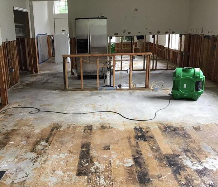 A living area that has had its floors and drywall removed to studs with a green air mover plugged in
